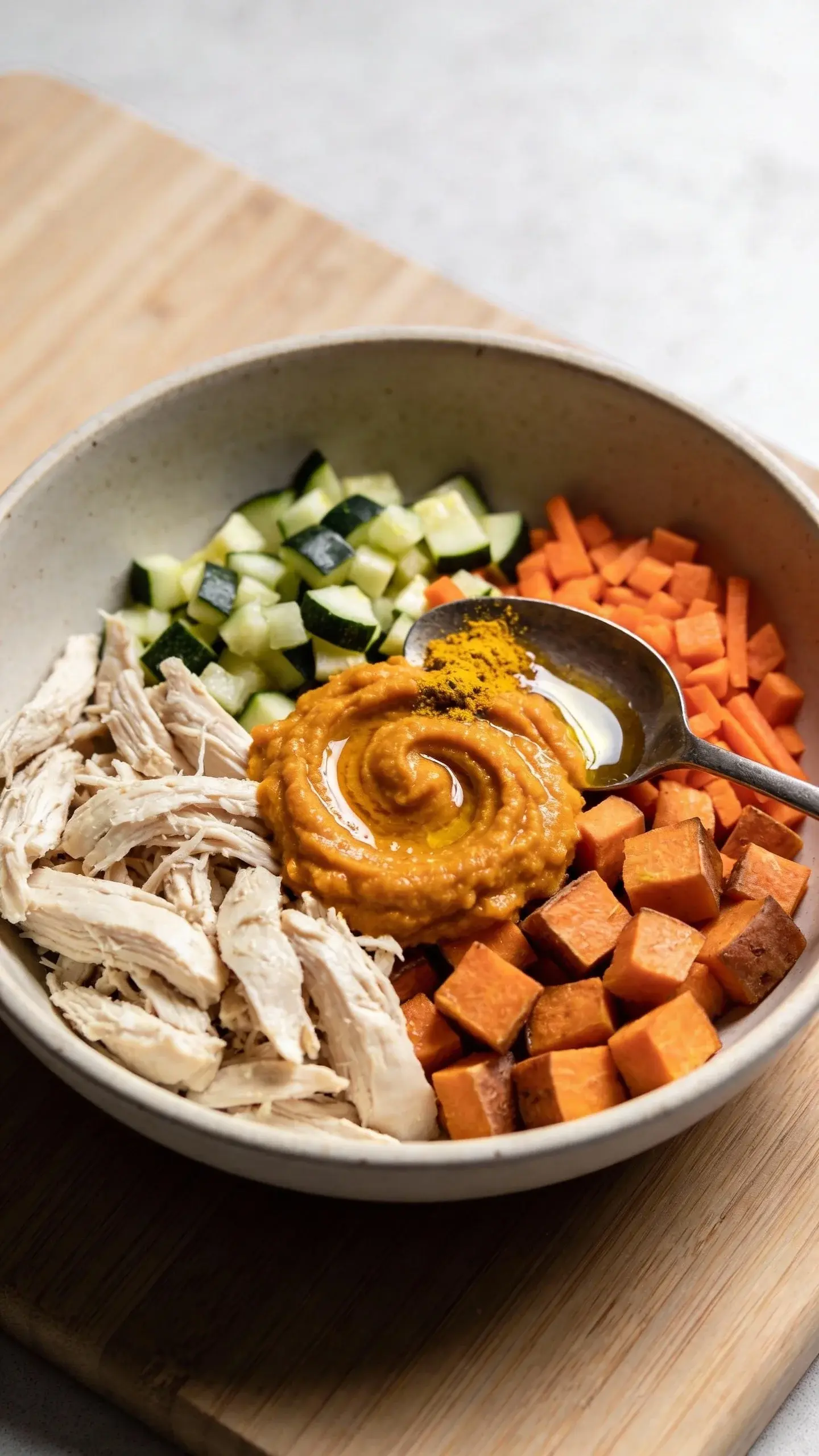 Close-up overhead shot of an allergy-friendly dog meal in a shallow ceramic bowl: shredded poached chicken breast, steamed diced sweet potato, finely chopped zucchini and carrots, a spoonful of mashed pumpkin swirled in, drizzled with a bit of olive oil, and a pinch of ground turmeric visible on top. Natural daylight, clean wooden countertop, soft neutral background, no text, high detail of textures and moisture.