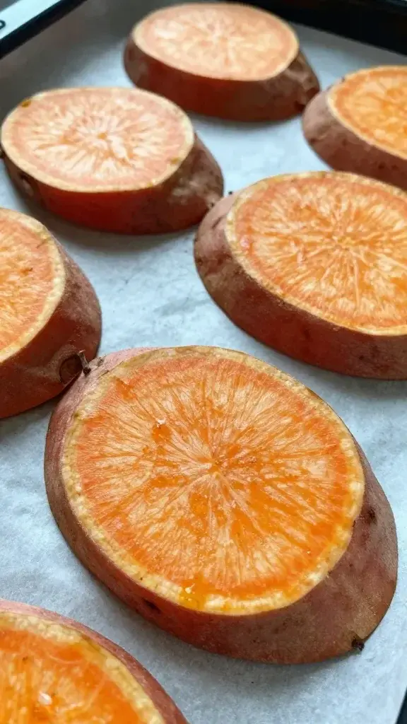 Close-up shot of sliced sweet potato rounds arranged on a parchment-lined baking sheet, with a light dusting of cinnamon visible, shallow depth of field focusing on the texture of the orange flesh and edges.